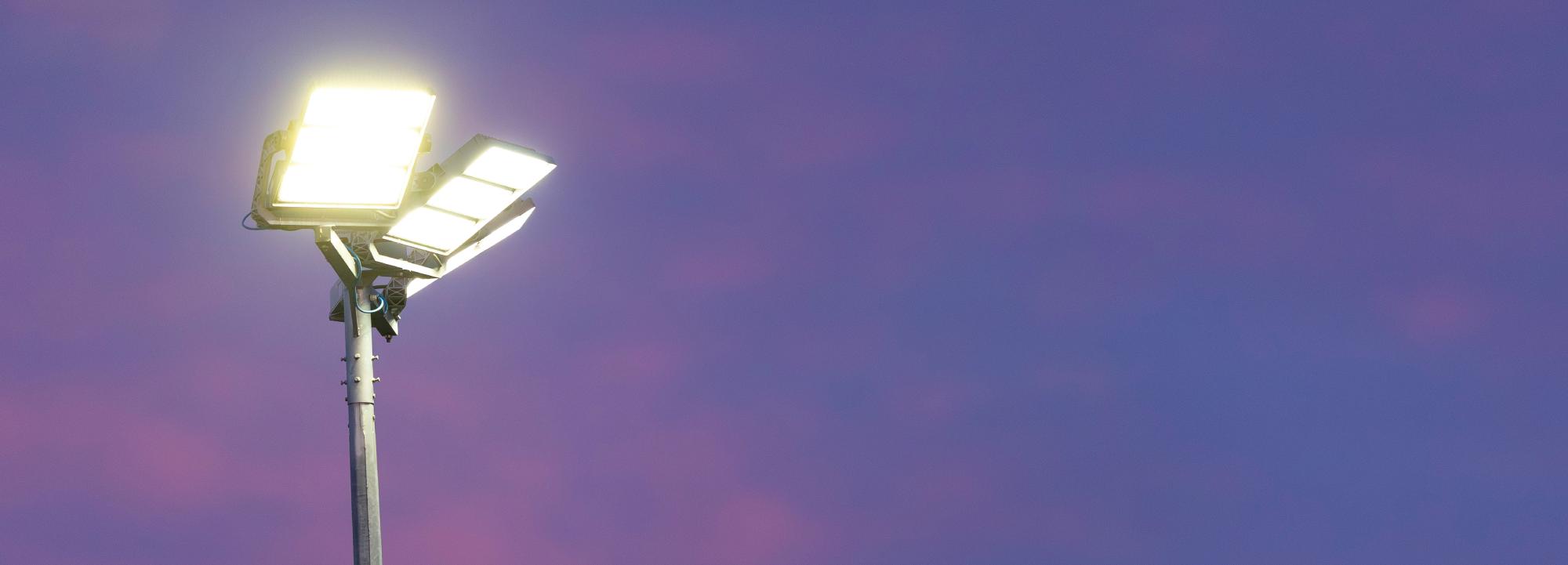 Two white floodlights attached to a tall metal pole against a blue and pink sky.