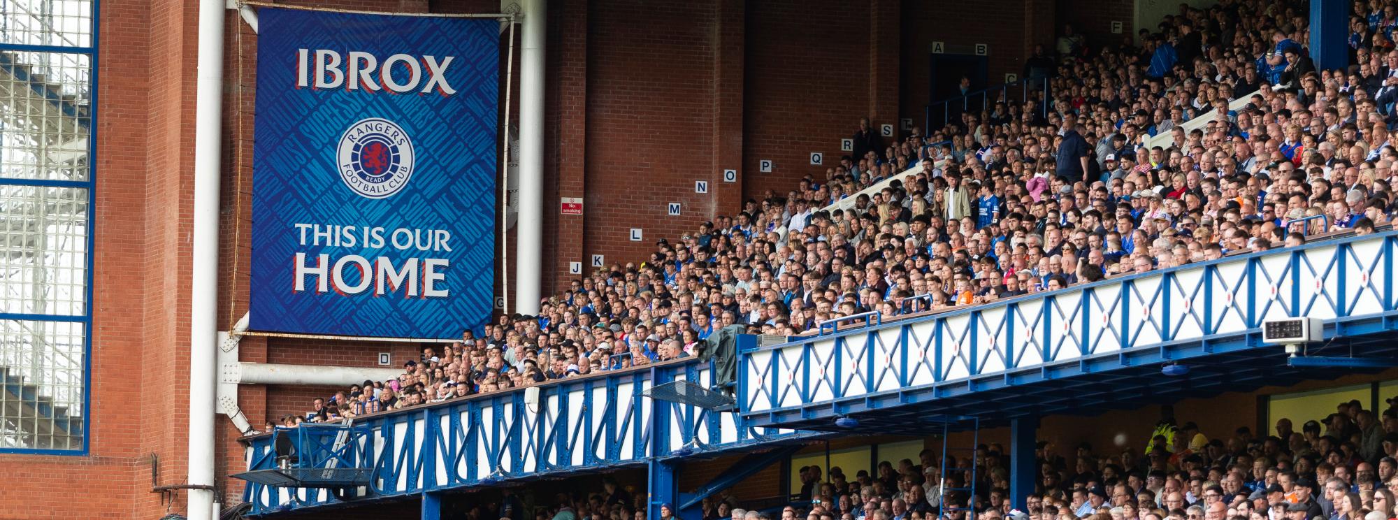 Banner reading "IBROX THIS IS OUR HOME" above packed stands at a football stadium
