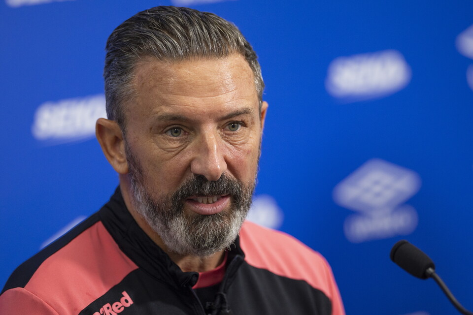 A man in a pink and black jacket speaks into a microphone.