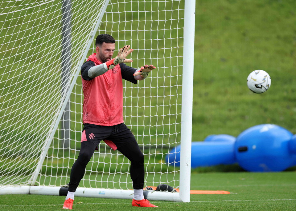 Footballer wearing red, black, and white training kit catching a ball with both hands.