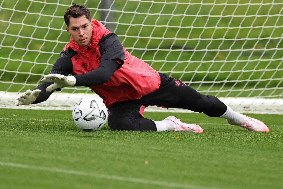 A goalkeeper in red and black training kit dives to catch a football.