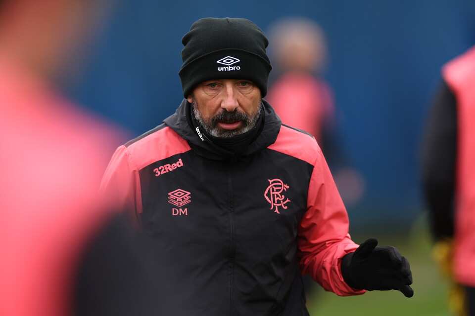 A man in a black and pink jacket with a red logo gives instructions on a football pitch.