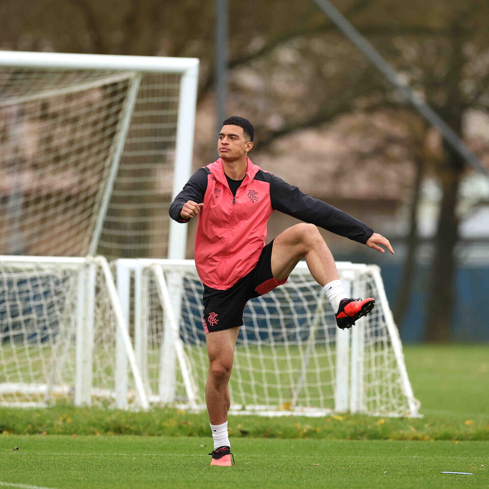 Footballer in red vest kicks ball toward goal during practice.