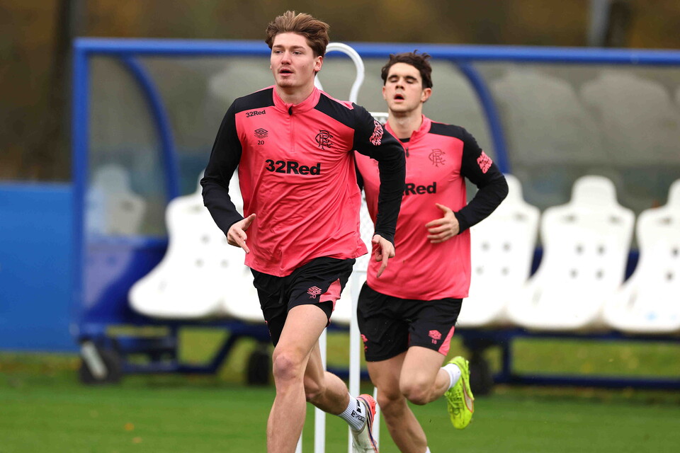 Two football players wearing pink shirts with red trims and black shorts are sprinting on a field.