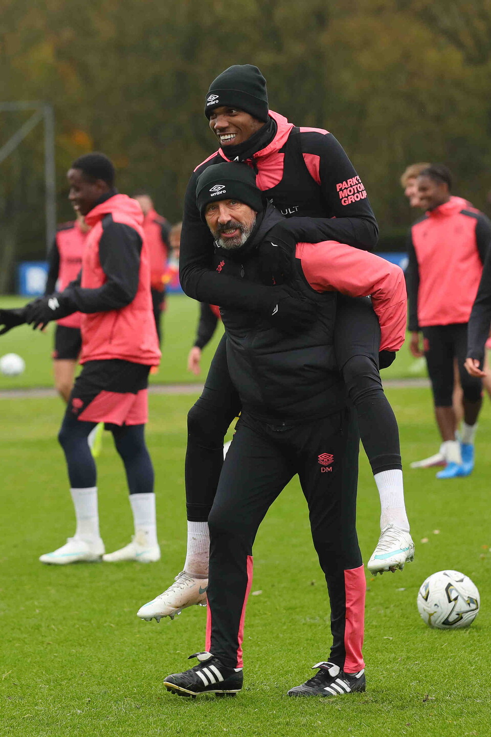 Football players in pink and black training suits and a coach in black with a white soccer ball nearby.