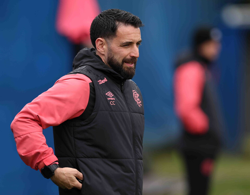 A football coach in a black and pink jacket looks to his side.