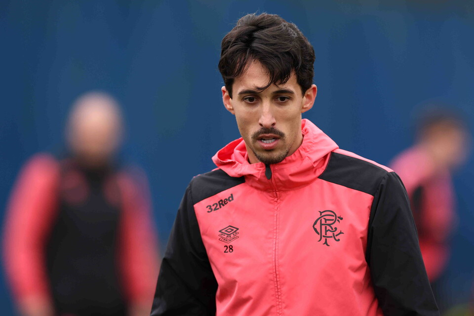 Footballer with a pink jacket walking on a field with blurred people in the background.