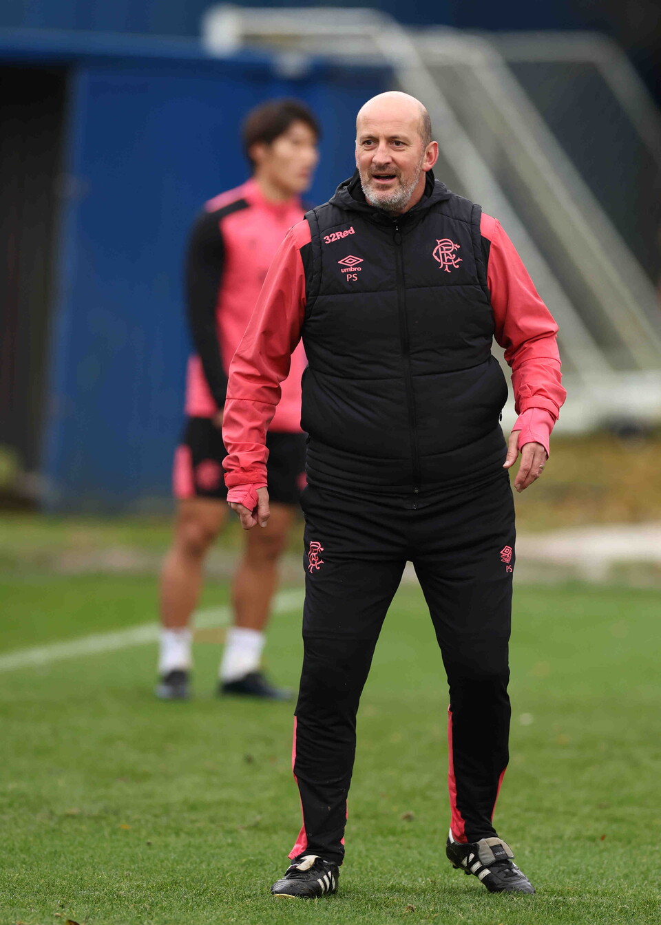 A coach in black and red attire on a green pitch with another player in pink training.