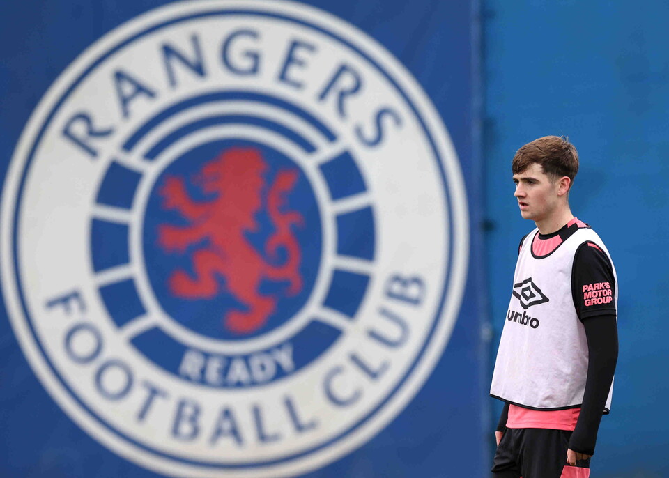 Rangers Football Club logo with a player in a white and black training shirt standing in front of it