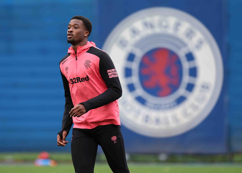 Player in pink and black training kit walking on football pitch with Rangers logo in background