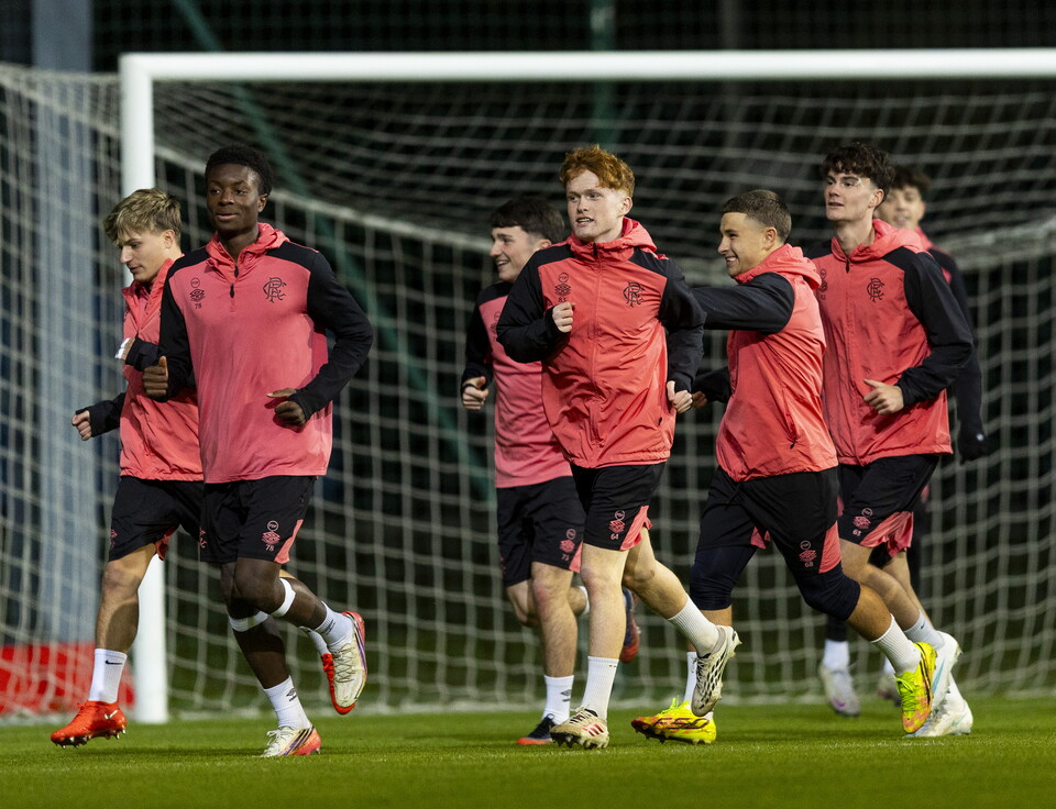 Seven players in red and black kits run on a football pitch, one leading with raised hands.