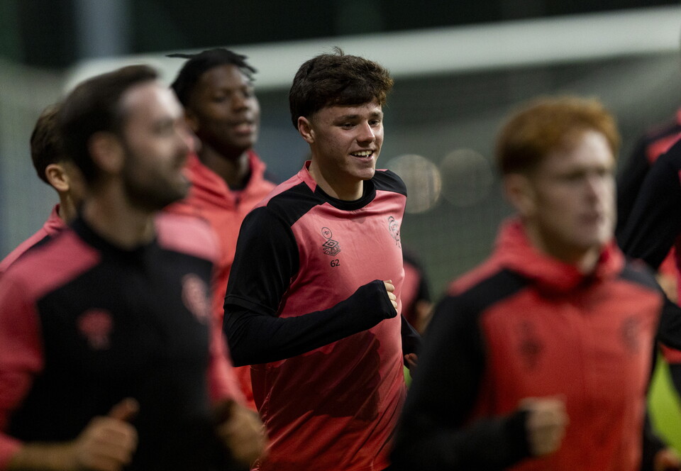 Six men wearing red and black training tops and shorts running on a football pitch.
