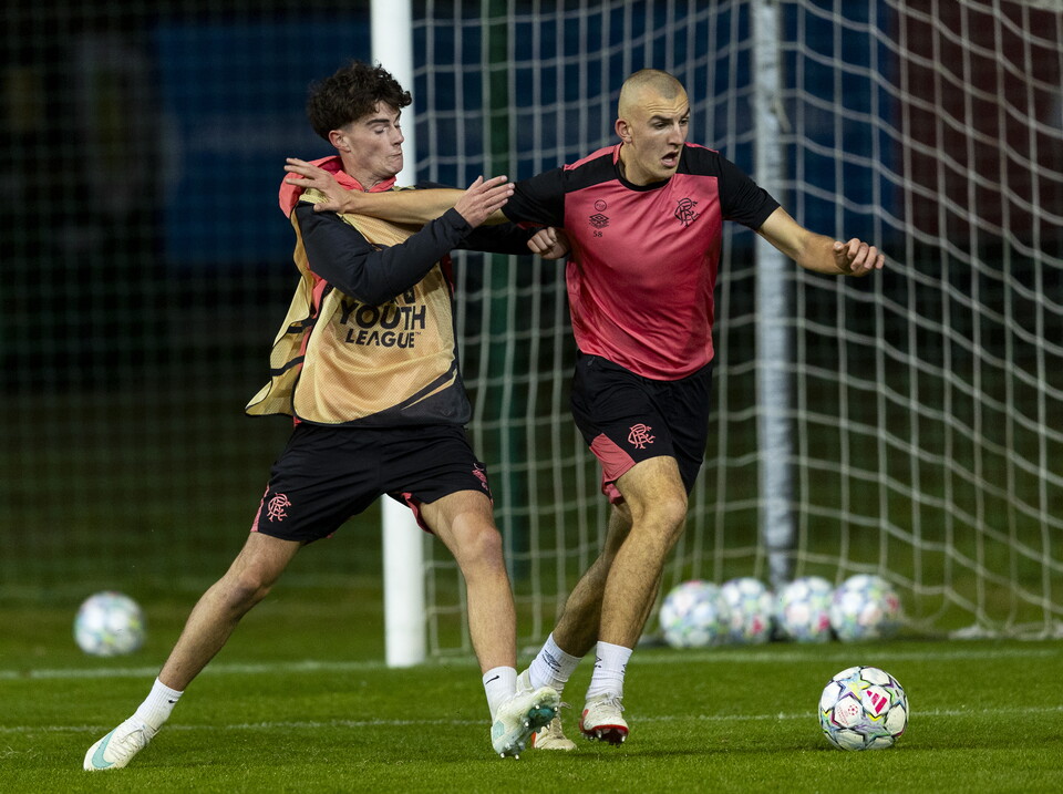 Two football players in yellow and red kits chase a soccer ball in front of a goal.