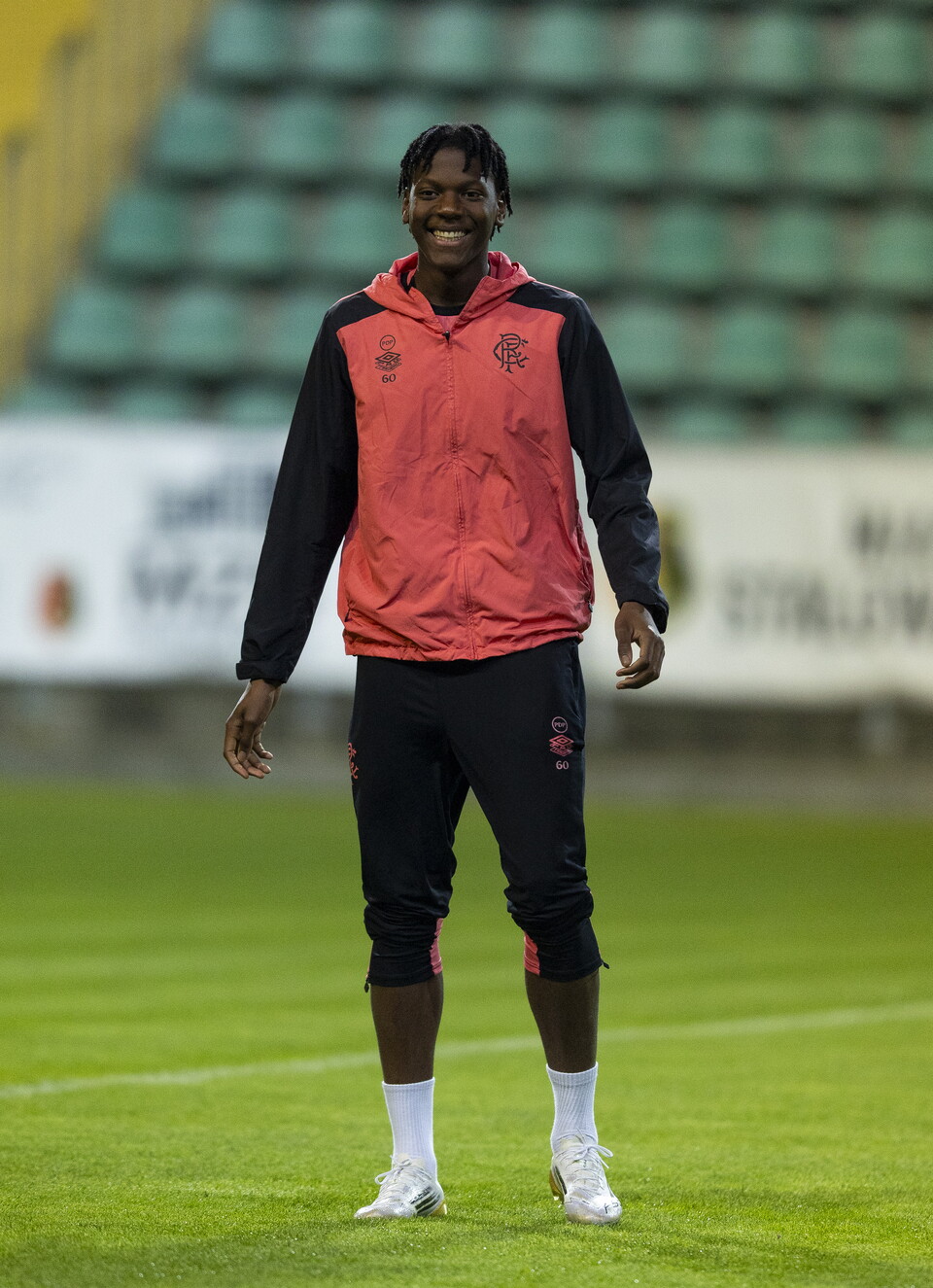 A footballer in red and black stands on a pitch with an empty grandstand behind.