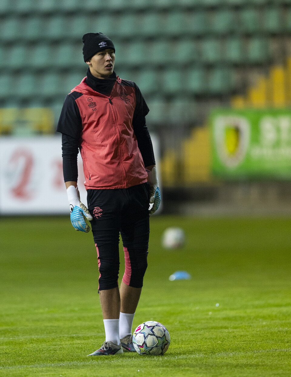 Goalkeeper standing in the field, wearing red, black and white training gear, gloves, and a blue hat.