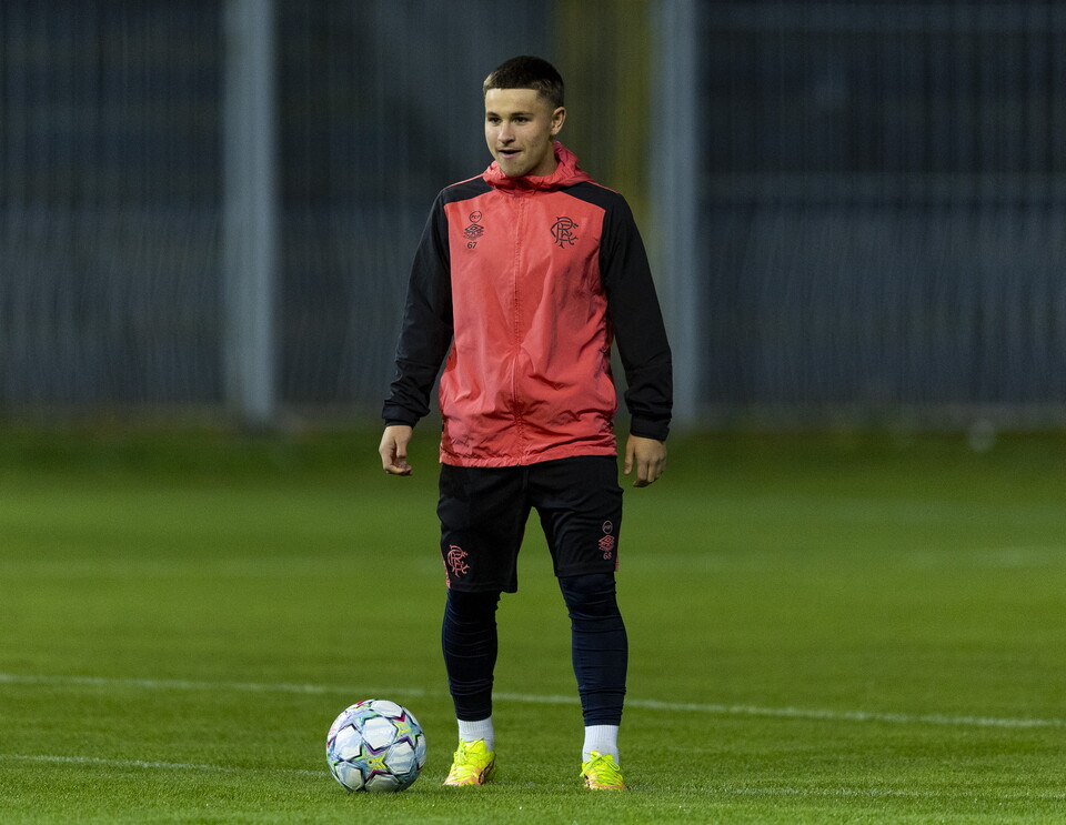 Football player in red and black training gear standing next to a soccer ball on a football pitch.