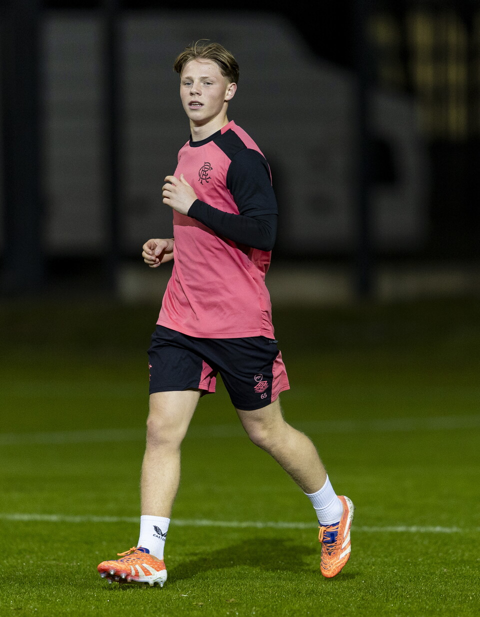 Footballer in pink shirt and black shorts running on grass field at night.