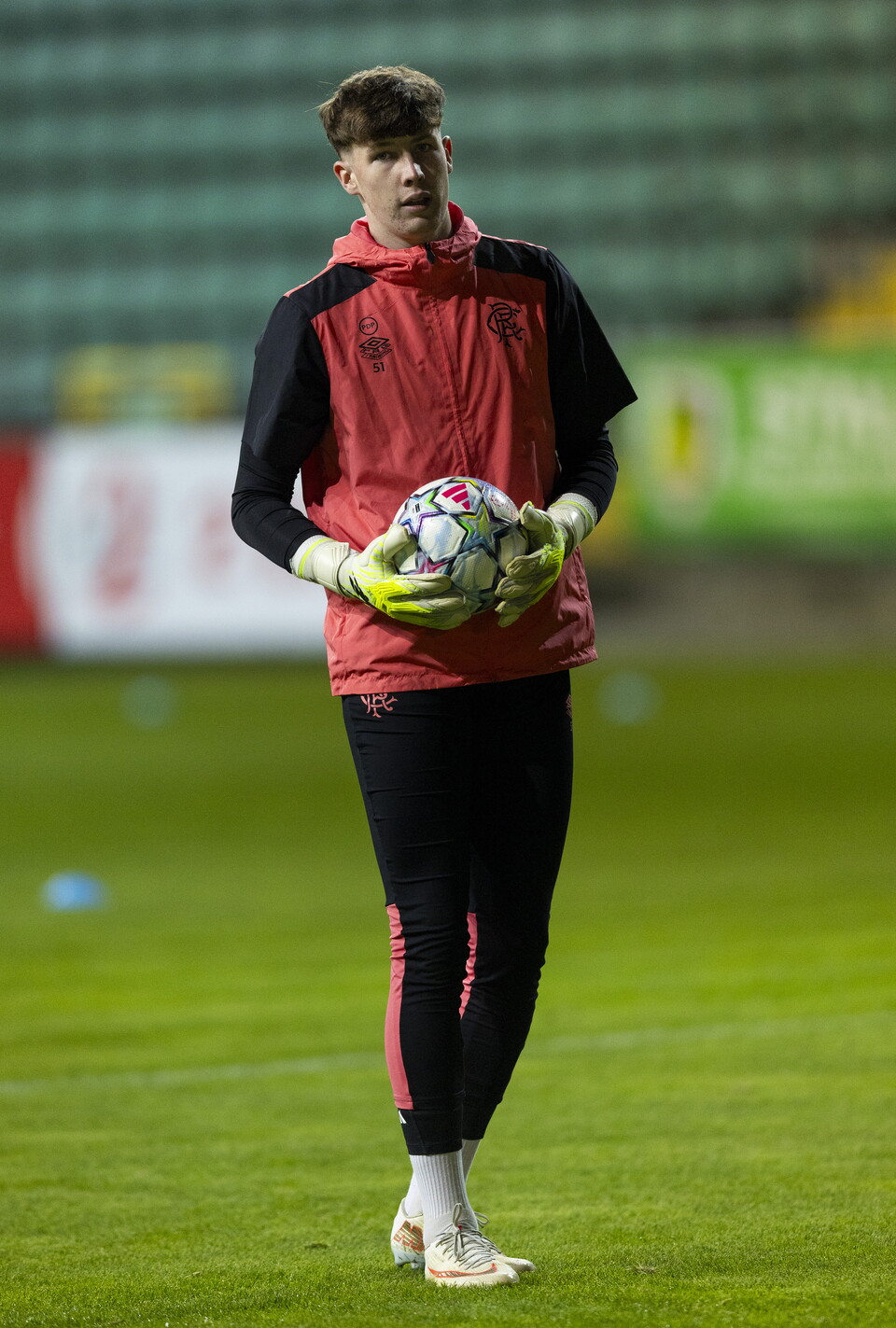 A man in red and black kit holding a football under his arm on a football pitch
