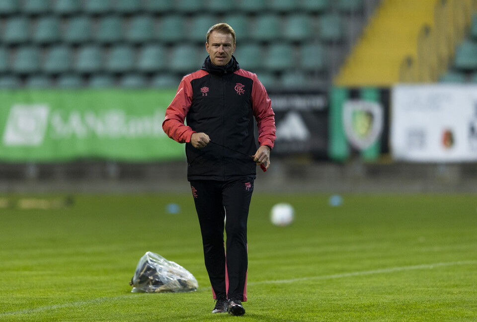 Coach in black and red outfit walks across the field with a bag in hand
