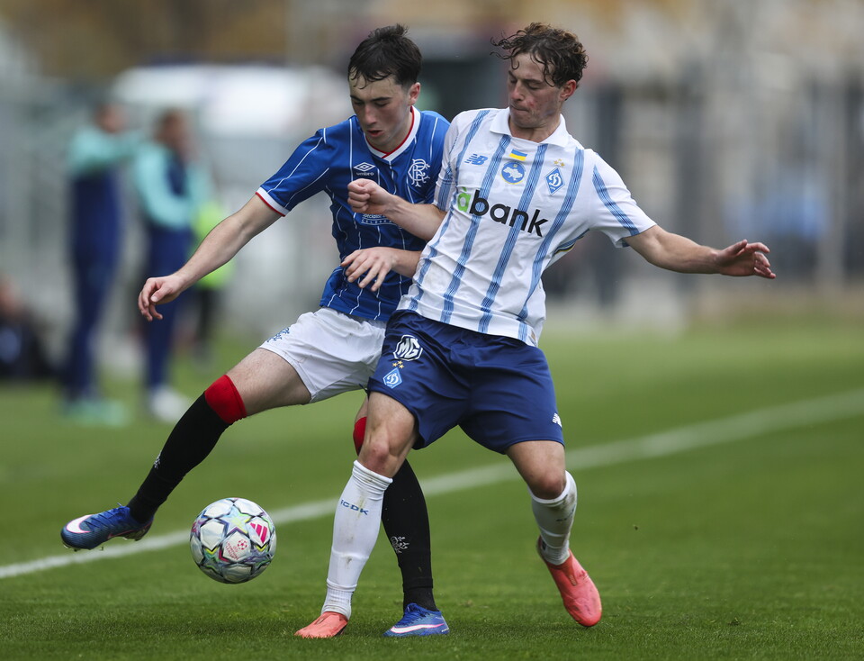 Two players in blue and white, red and white jerseys challenge for a soccer ball on a green field.
