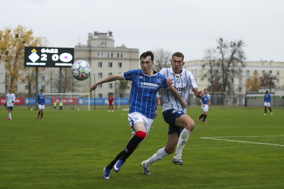 Two football players chase a ball mid-air in a field with a building in the background.