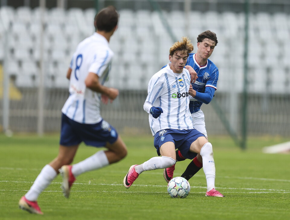 Three football players, one in white and blue, one in blue, and one in white, compete for the ball.