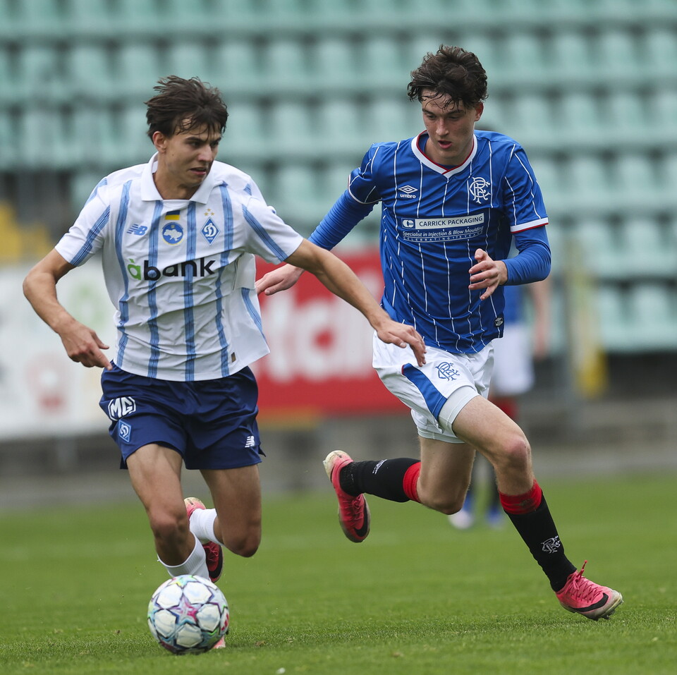 Two players on a football pitch; one chases the ball in a white shirt, while the other chases in blue.