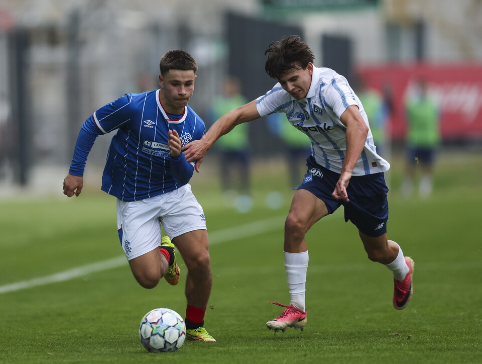 Two players in blue and white kits chase a soccer ball on a field.