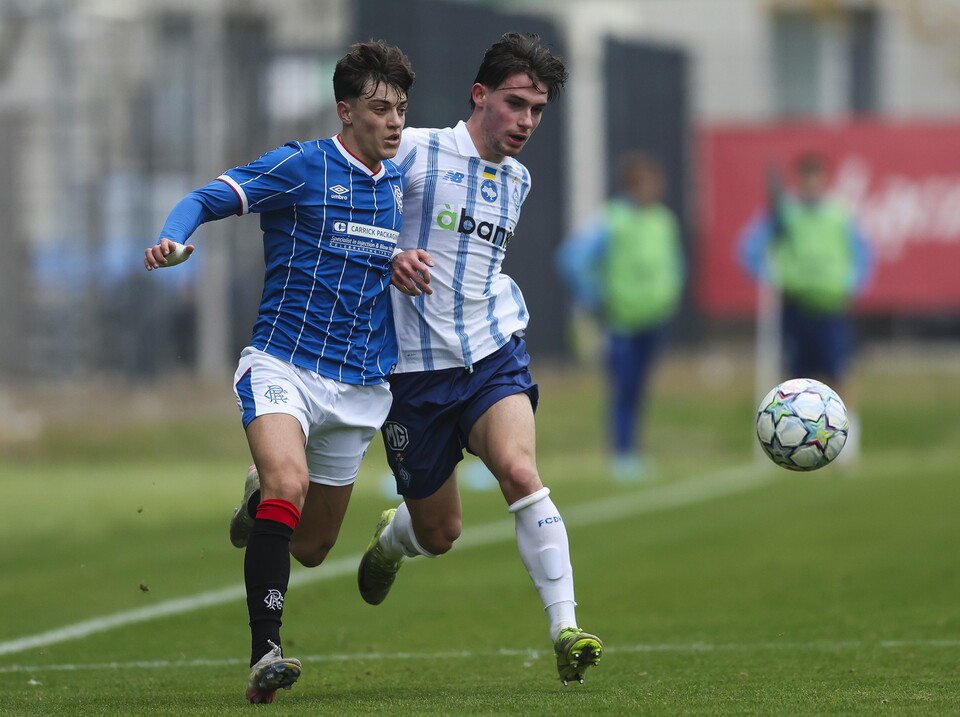 Two soccer players in blue and white jerseys chase a soccer ball on a field.