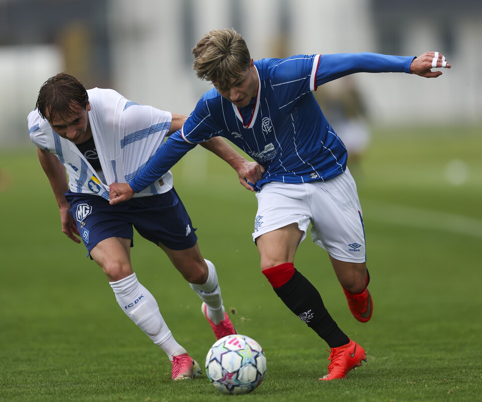 Two soccer players, one in a blue jersey, the other in a white jersey, contest a soccer ball.