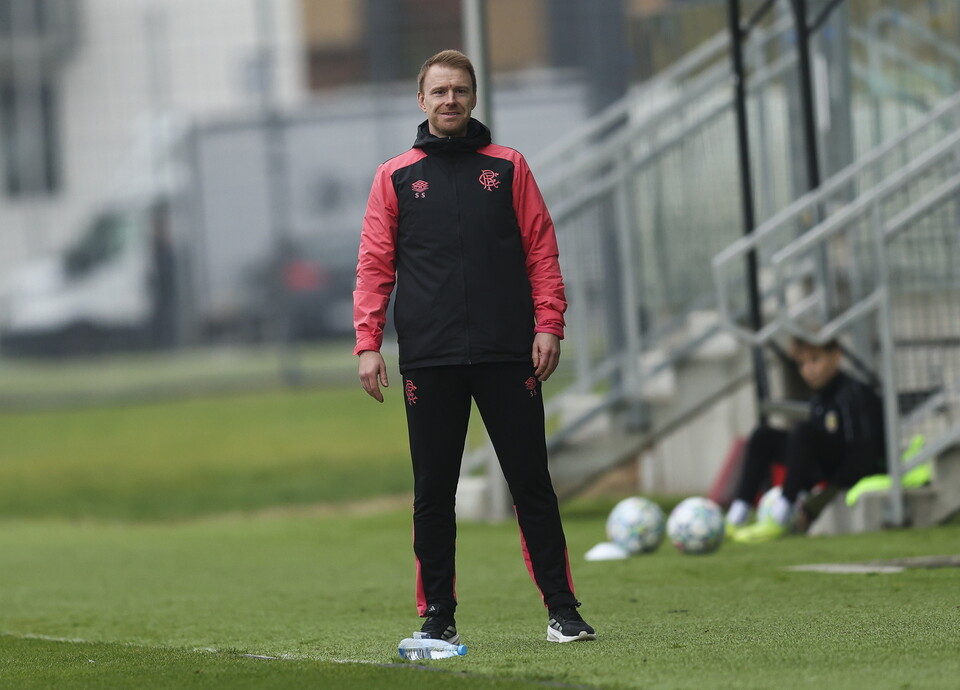Man in red and black tracksuit watches two players on grass pitch.
