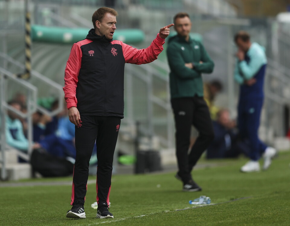 Coach gesturing and shouting on a football pitch with two men in the background