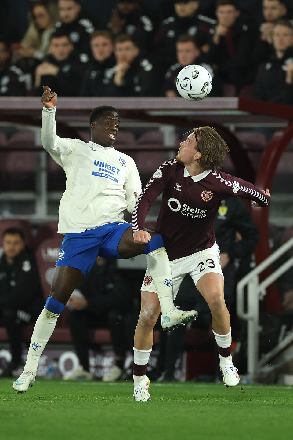 Two football players jump to head a ball in mid air; one in a white shirt, blue shorts and white socks, the other in a maroon shirt, maroon shorts and white socks.