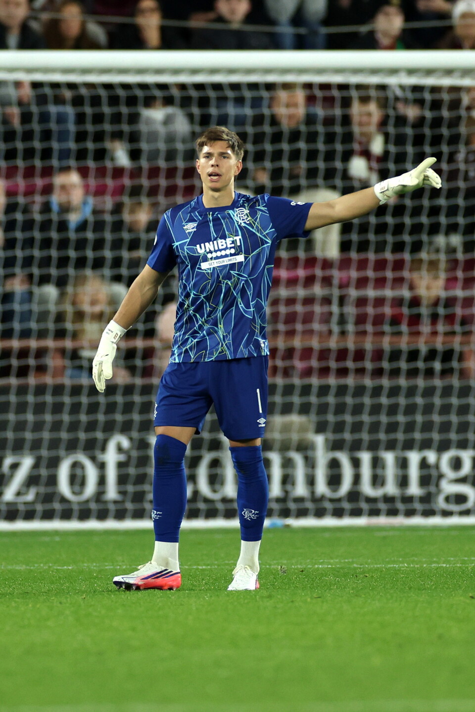 Football player wearing blue kit stands on pitch, arms outstretched, crowd behind him