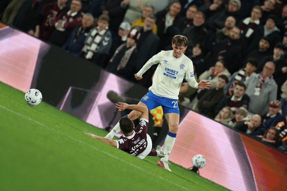 Two players on a football field. One player in white kicks the ball while another in maroon tries to block.