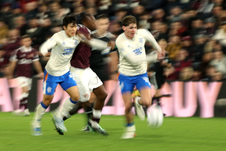 Footballers in white and blue kits chase a football in an open stadium.