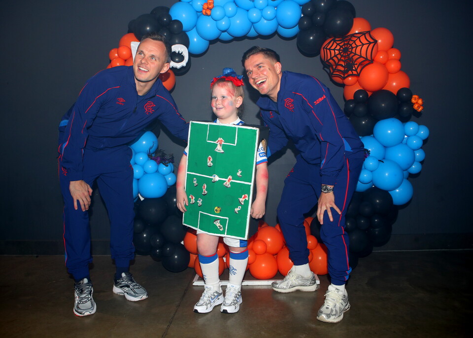 Three men and a girl pose behind a balloon arch, girl holds a football pitch board.