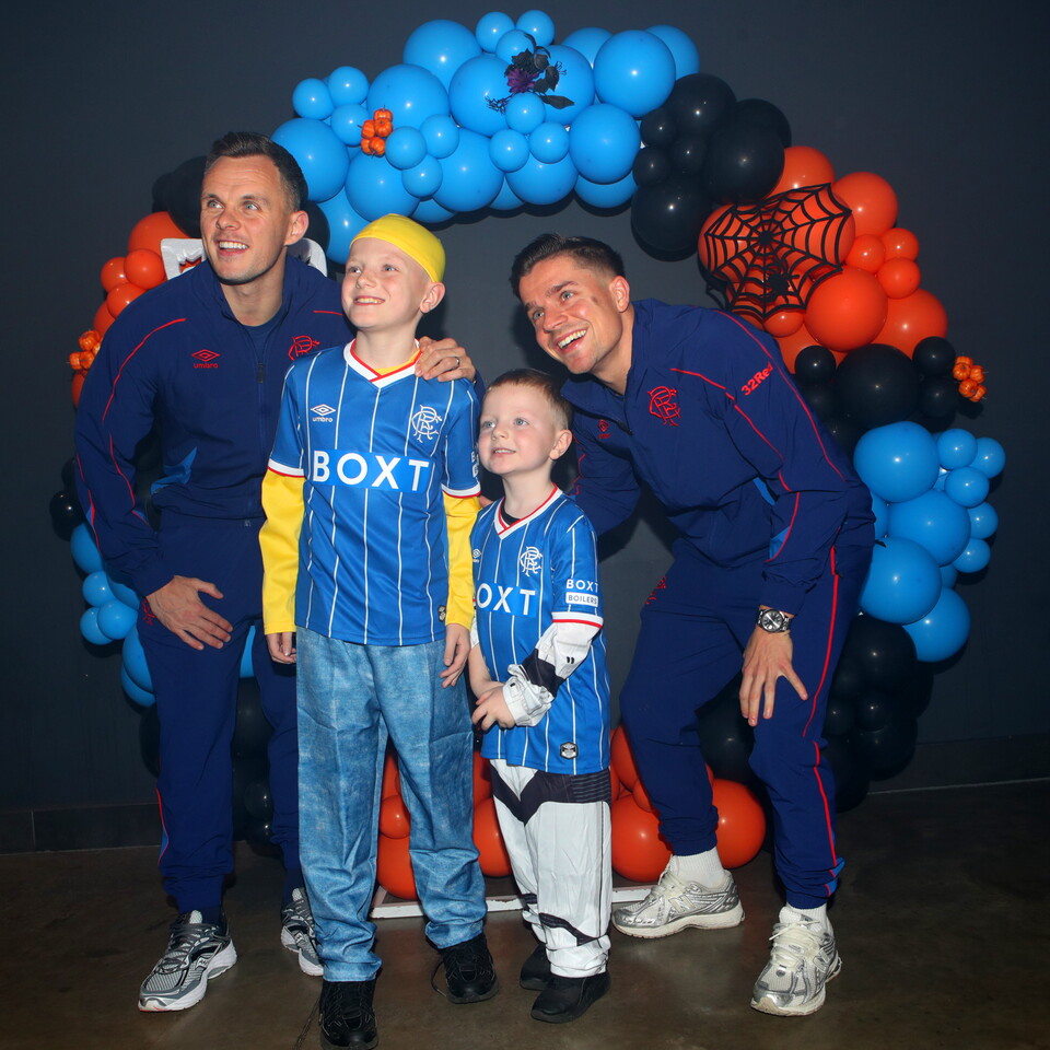 Two men and two children pose for a photo in front of an orange and blue balloon arch.