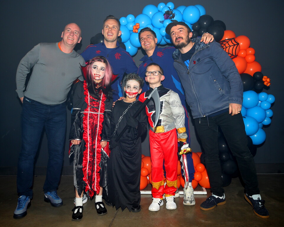 Seven people dressed in Halloween costumes posing in front of a balloon arch.