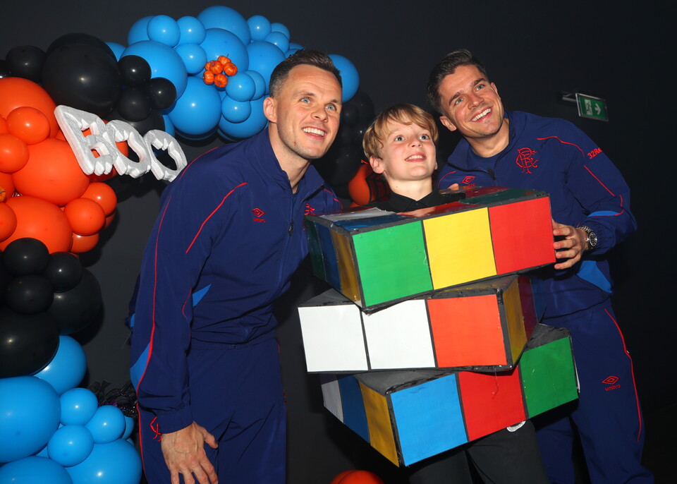 Three men in blue jumpsuits, one holding a giant Rubik's cube, pose in front of a balloon backdrop.
