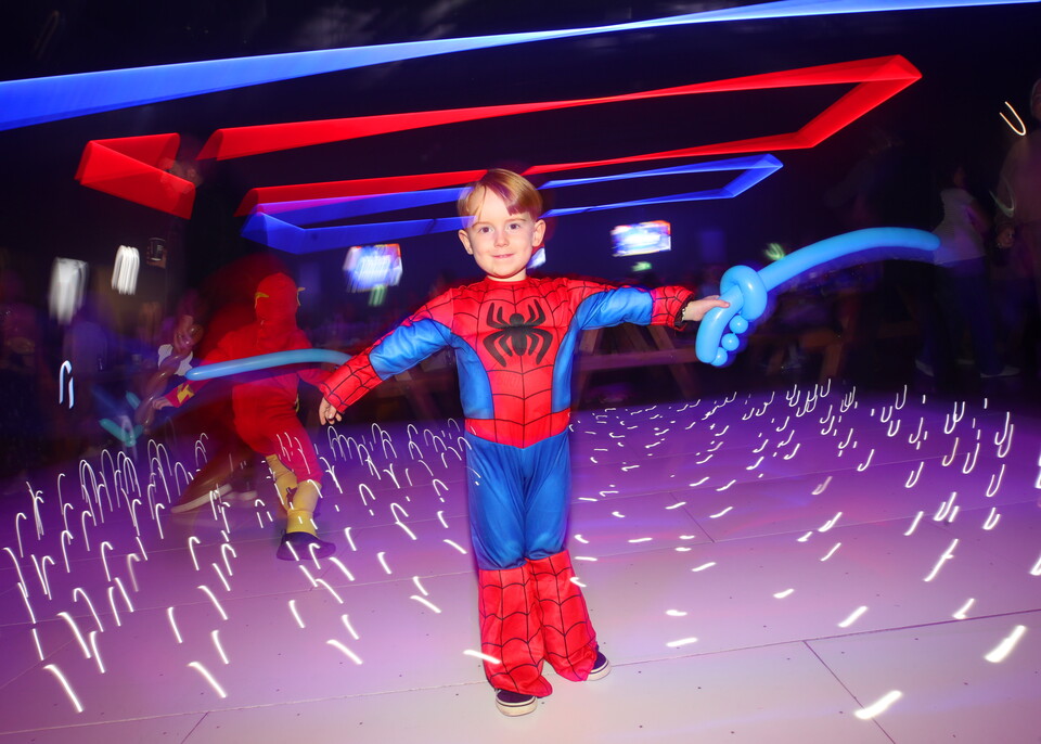 A child dressed in a Spiderman costume holds a balloon on a dance floor with glowing purple lights