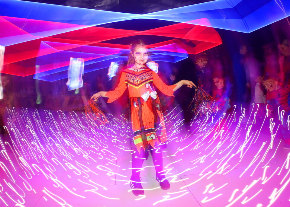 Child in orange dress and purple boots performing on a stage with colorful lights.