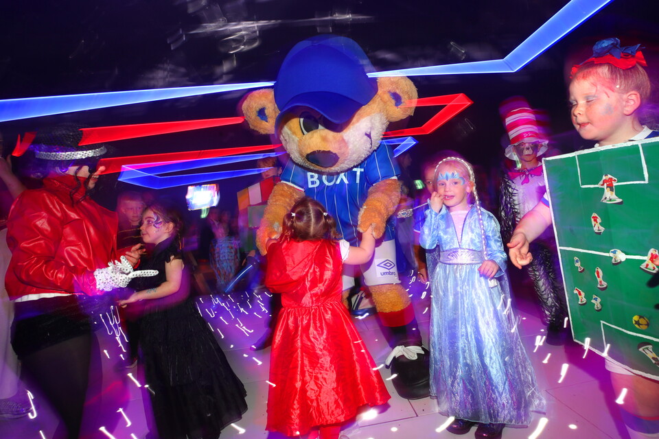 Kids in costumes dance around a mascot dressed in a football shirt at a party.