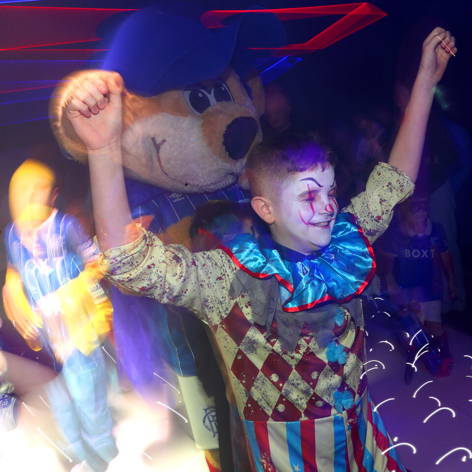 A child dressed as a clown pumps fists in the air next to a large mascot.