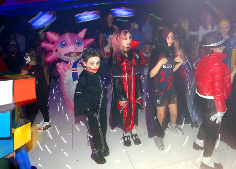 Children in costumes on a dance floor with a mascot behind them.