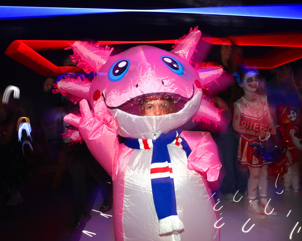 Child in pink dragon mascot waving at people in a hall with red and blue lighting.