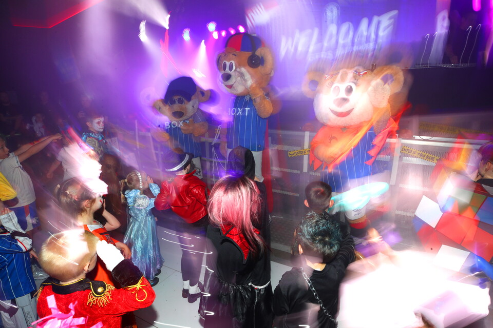 Several children in costumes dance in a nightclub, large mascots are in the background.