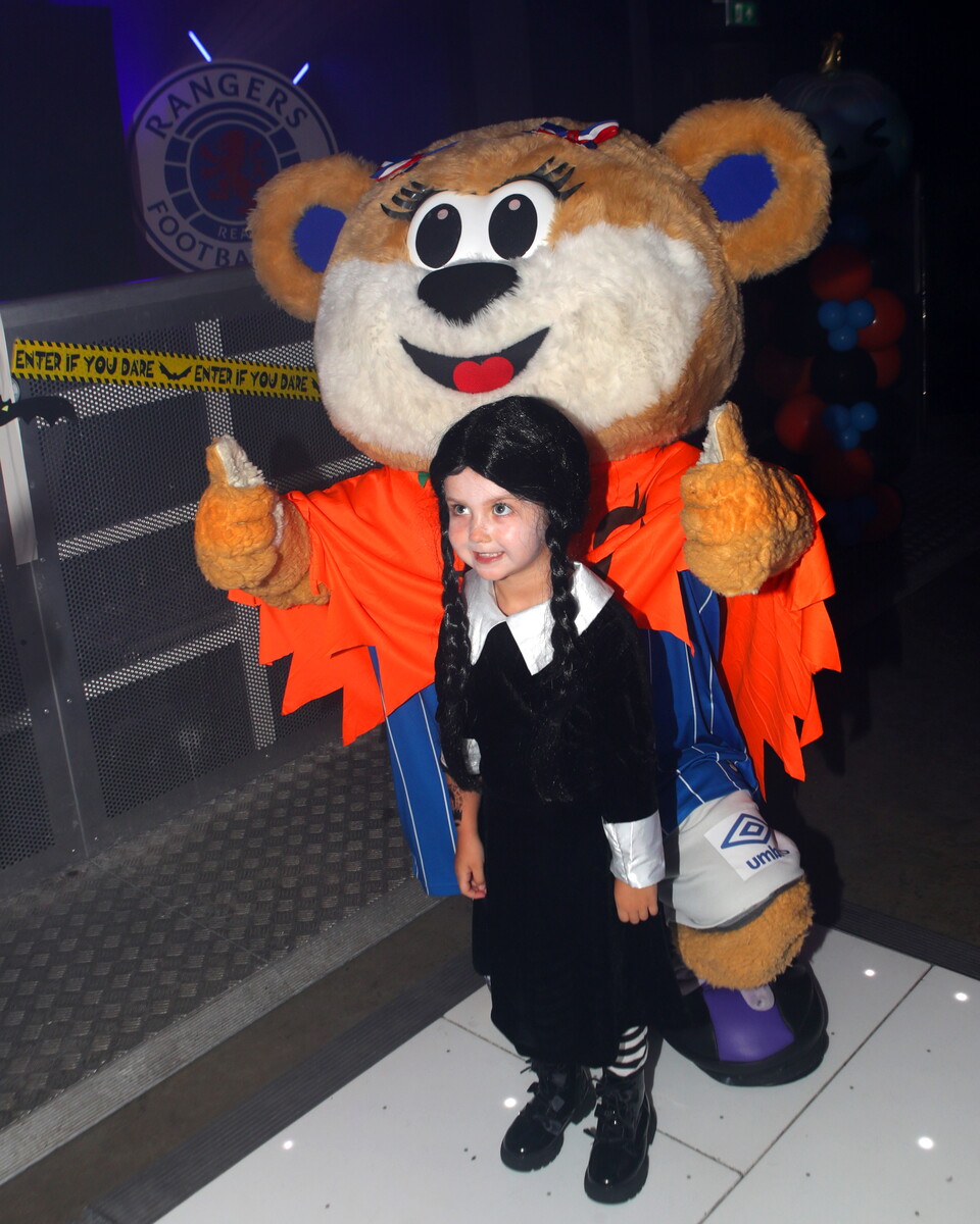 A small girl poses with a giant Teddy mascot at an indoor event.