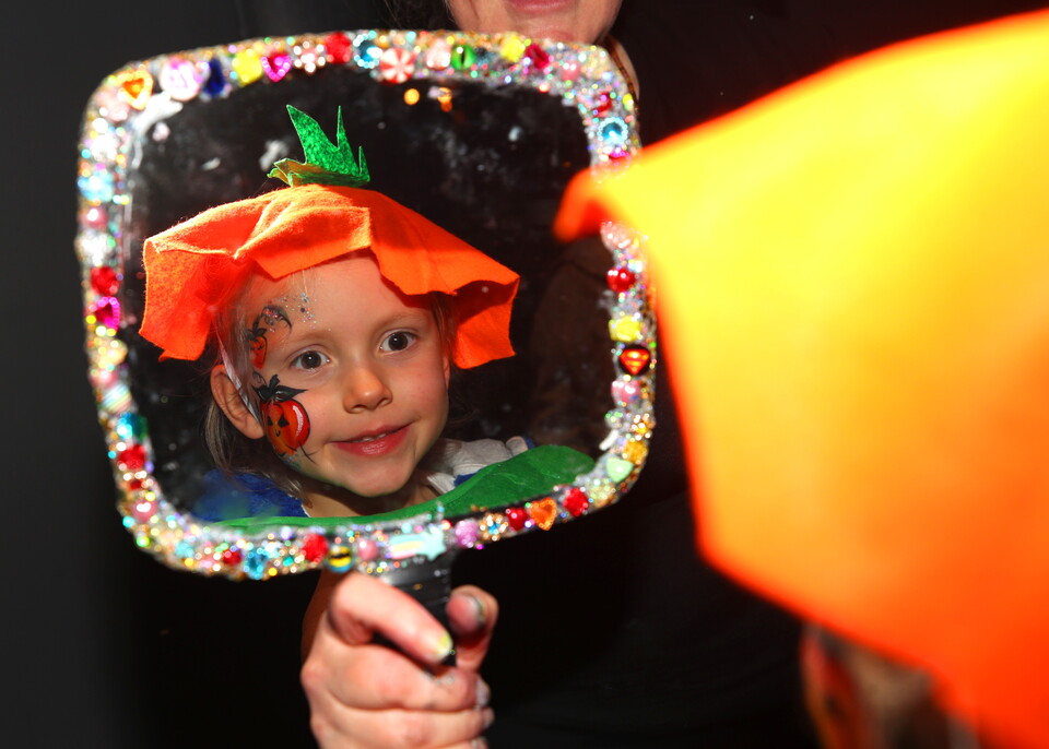 Young girl in pumpkin hat with red cheeks and ladybug face paint looking in mirror.