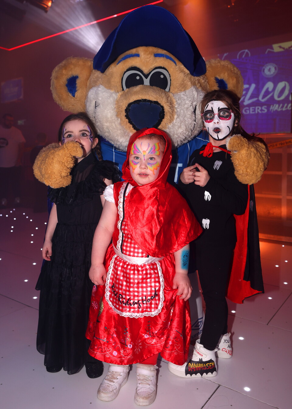 Three children dressed up as a witch, vampire and Little Red Riding Hood standing with a large mascot bear